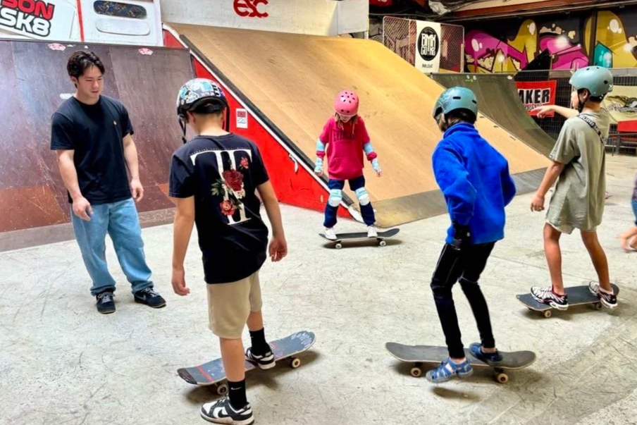 Four children wearing helmets skateboarding indoors at a skate park, with a young adult supervising.