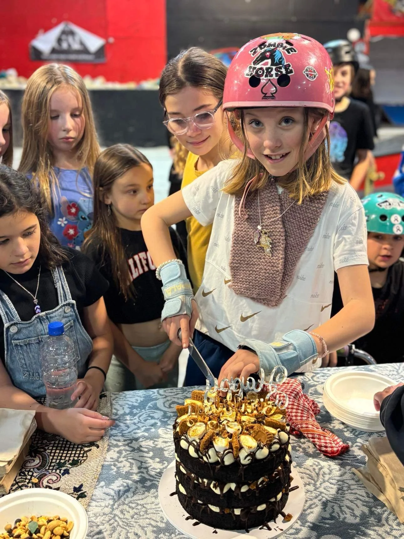 A young girl wearing a pink helmet with 'Zombie Horse' written on it, cutting a birthday cake with birthday decorations, surrounded by children at a celebration.