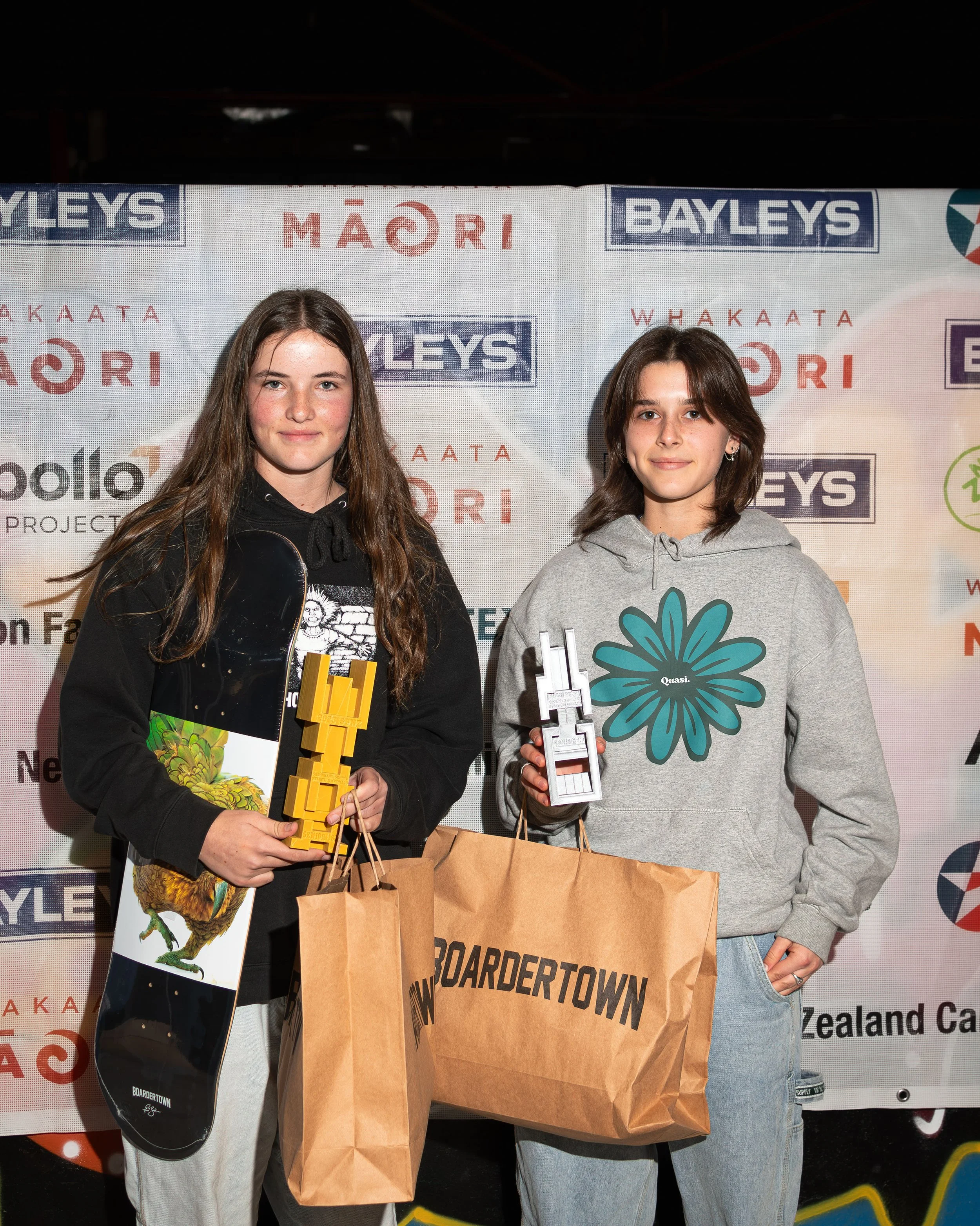 Two people holding skateboards and trophies, standing in front of a backdrop with logos, holding shopping bags labeled 'Boardertown.'