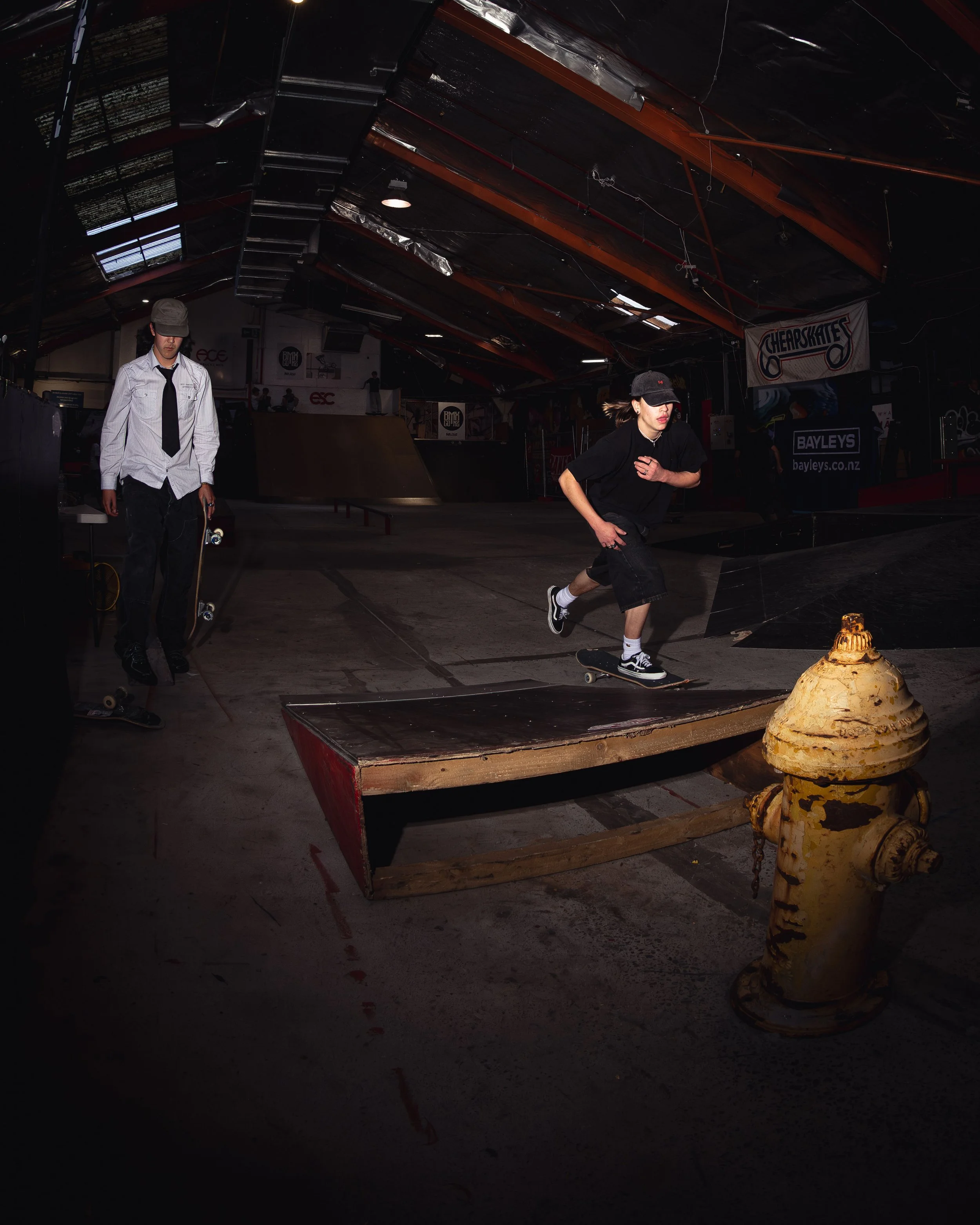 Indoor skate park with two people; one walking with skateboard, the other jumping over a ramp.