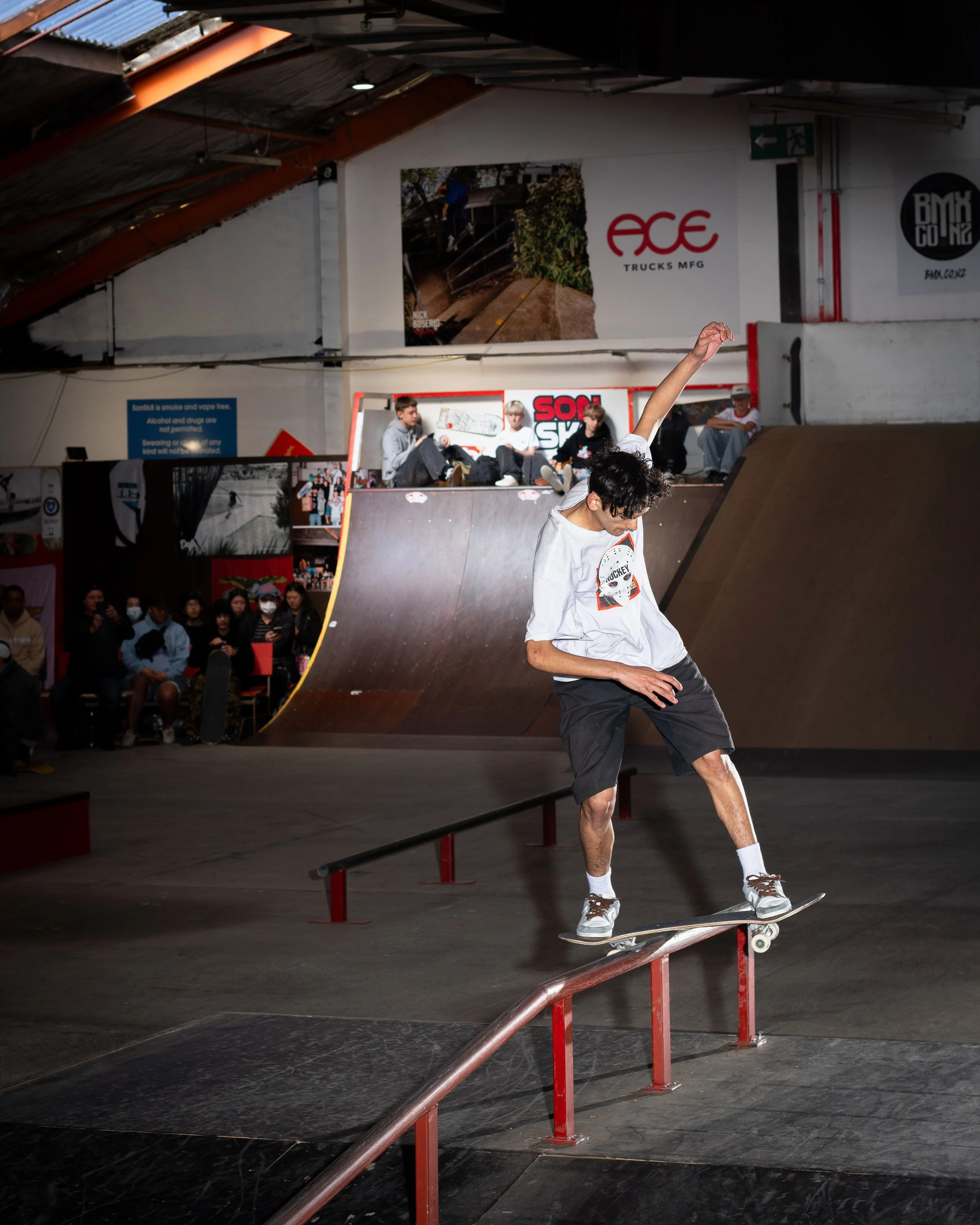Skateboarder performing a grind trick on a rail in an indoor skatepark with spectators in the background.