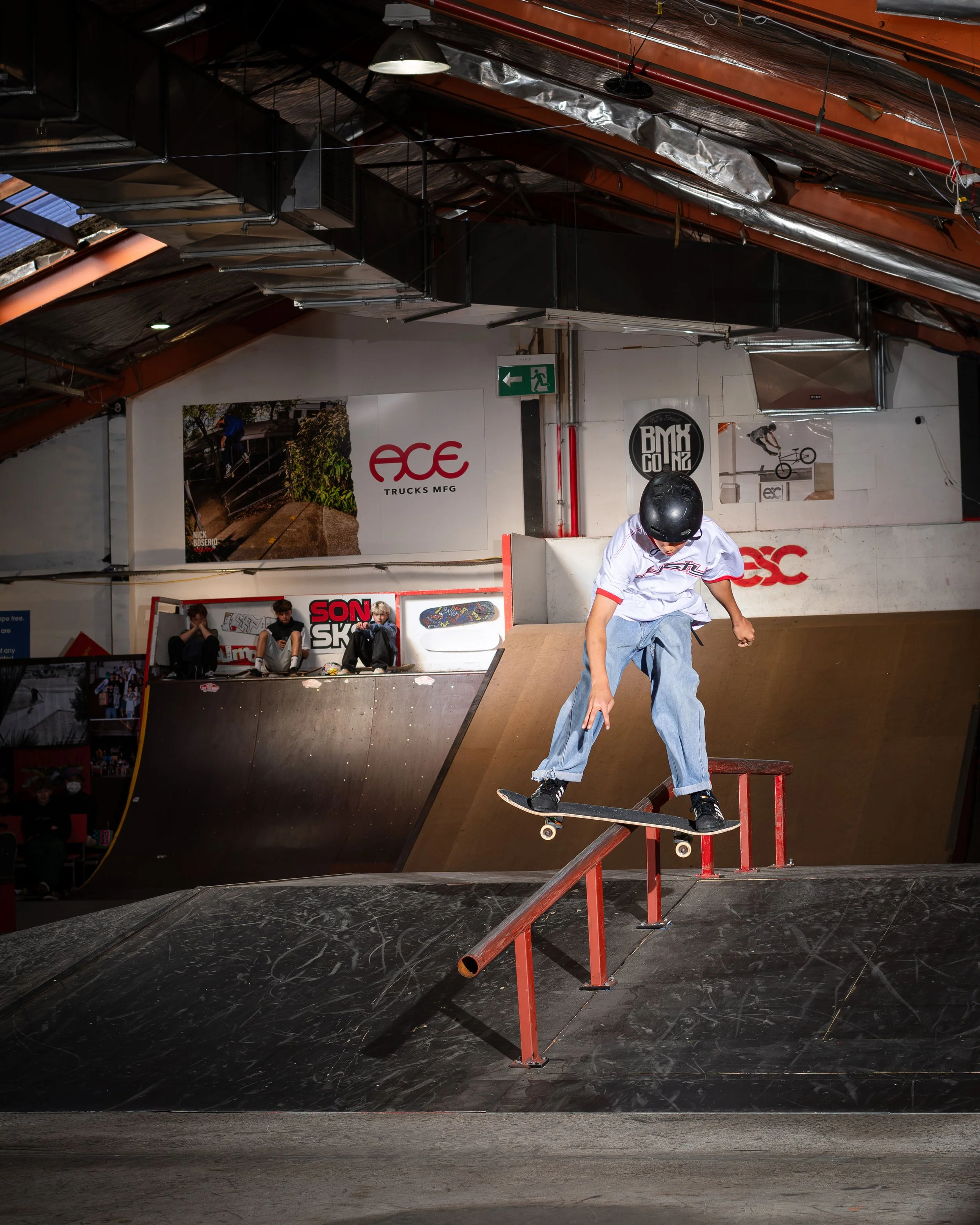 A person wearing a helmet skateboarding on an indoor ramp, performing a trick over a rail with spectators watching in the background.