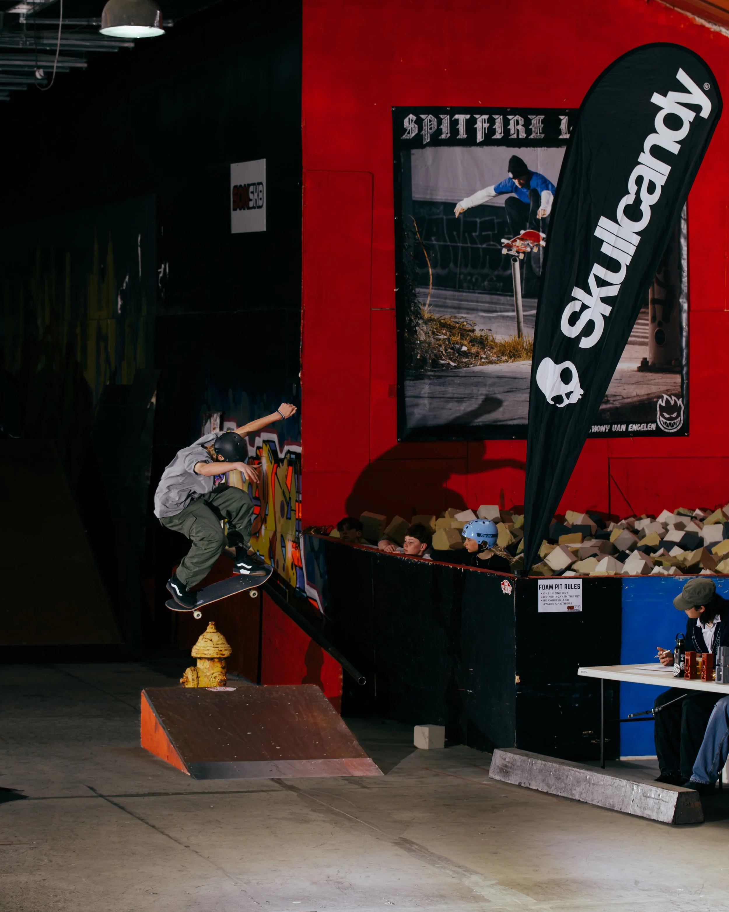 Indoor skatepark scene with a skateboarder performing an ollie over a yellow fire hydrant. There is a Skullcandy banner and a Spitfire poster in the background. Spectators watch the trick near a foam pit.