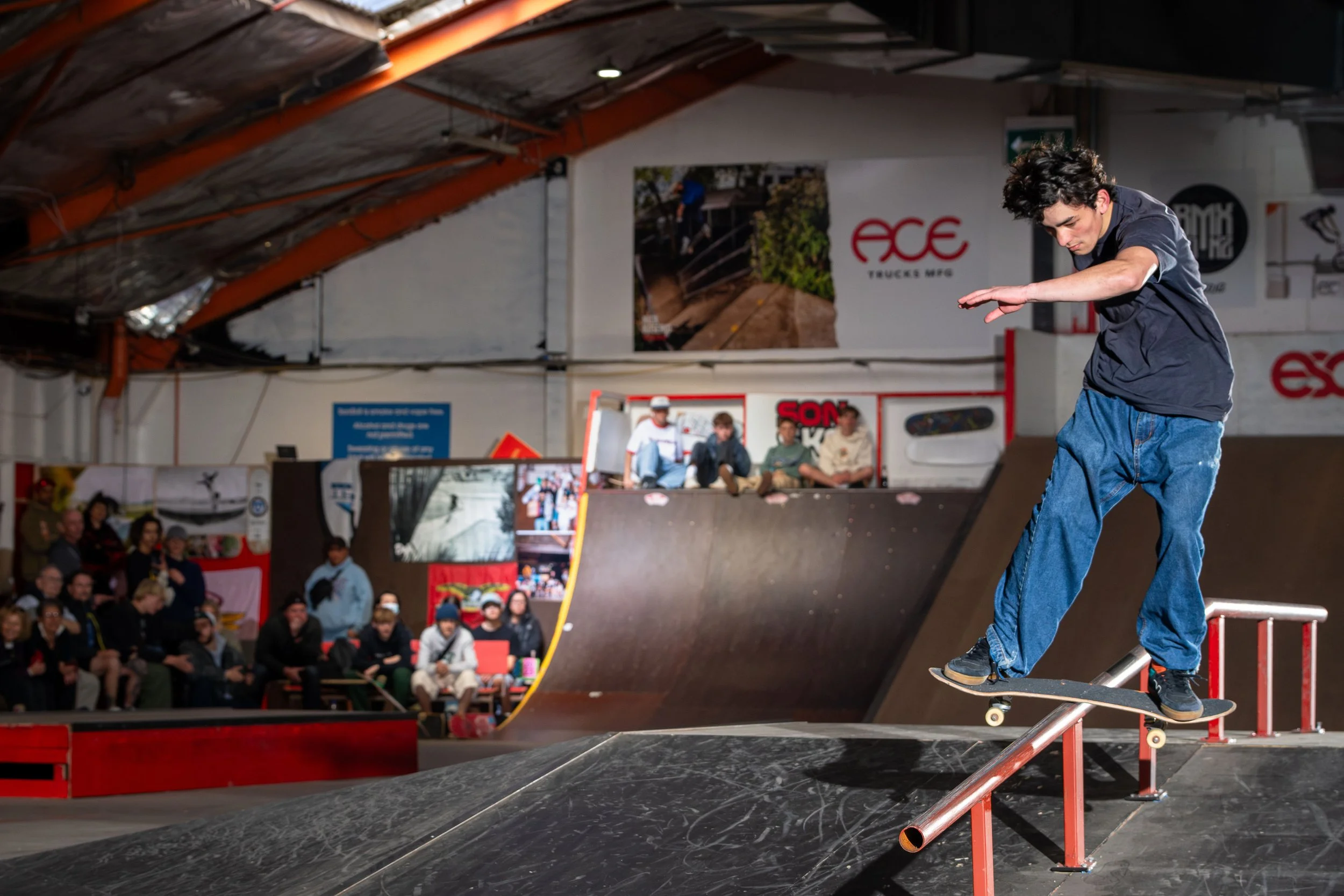 Skateboarder grinding on a rail indoors with spectators watching in a skatepark.