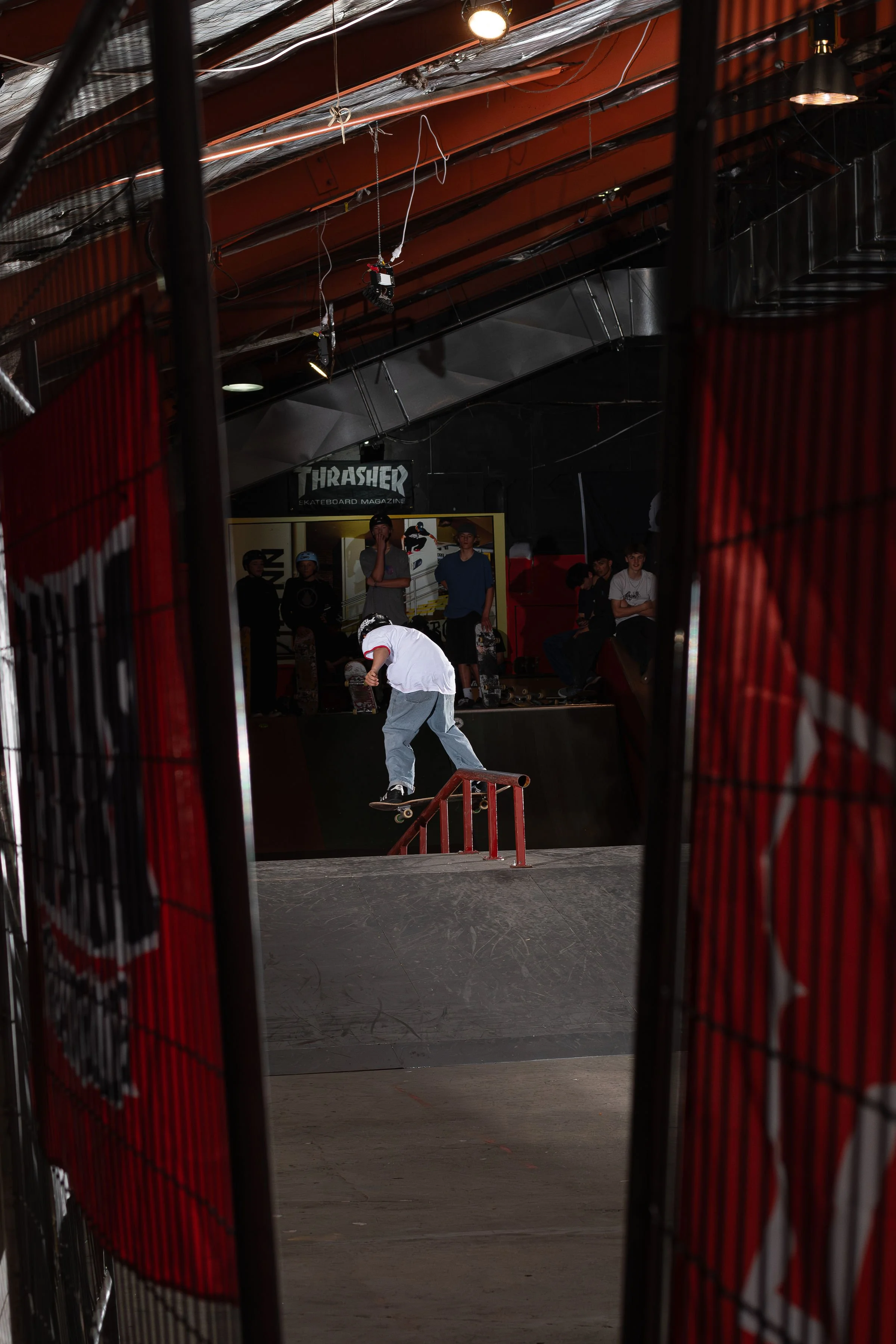Skateboarder performing a trick on a rail in an indoor skate park, surrounded by spectators. The environment includes a ramp, beams, and industrial lighting, with a "Thrasher" sign in the background.