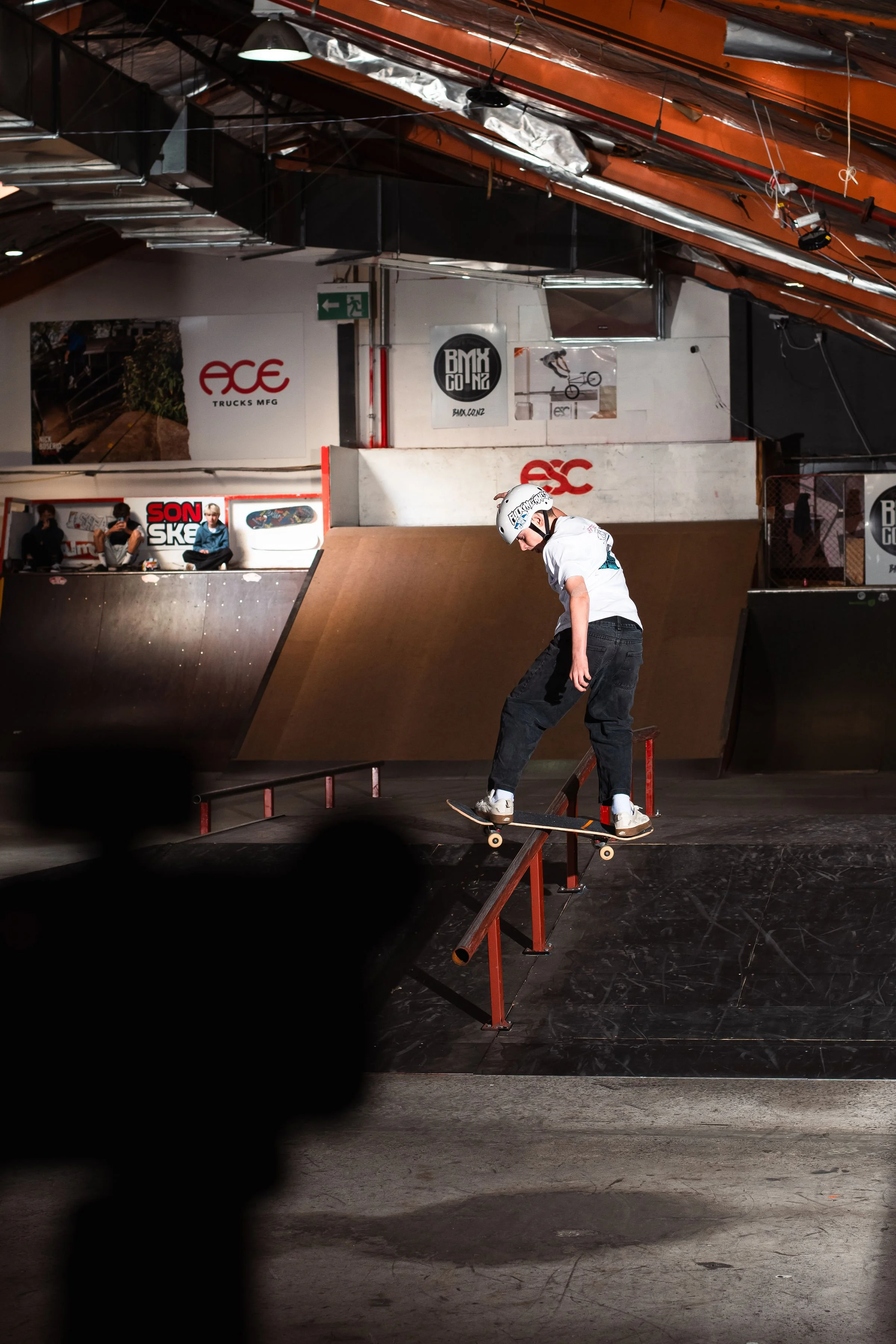 Person skateboarding on a rail in an indoor skate park, wearing a helmet and casual clothing. The park features ramps and posters on the walls.