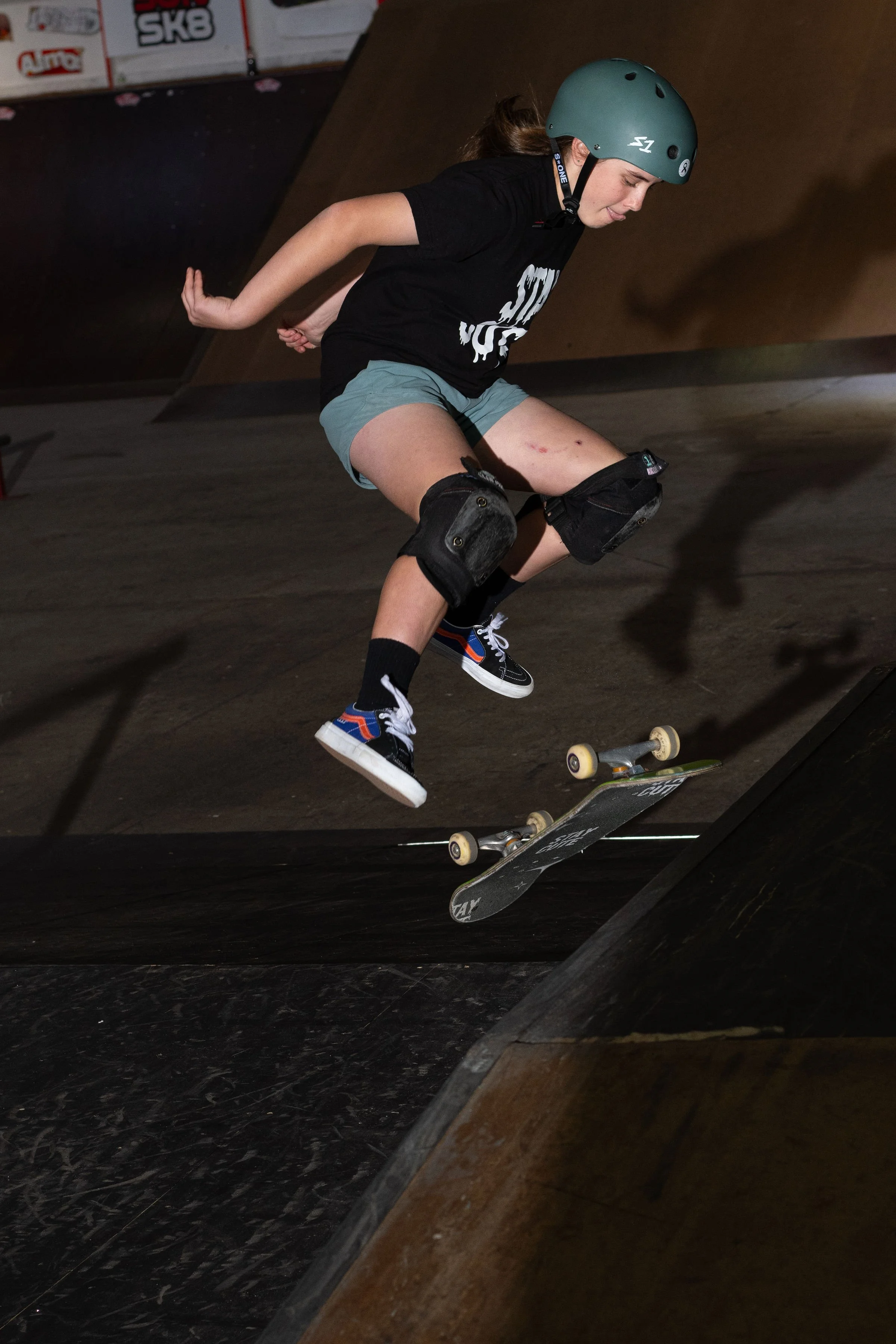 A person performing a skateboard trick in an indoor skate park, wearing a helmet and knee pads.