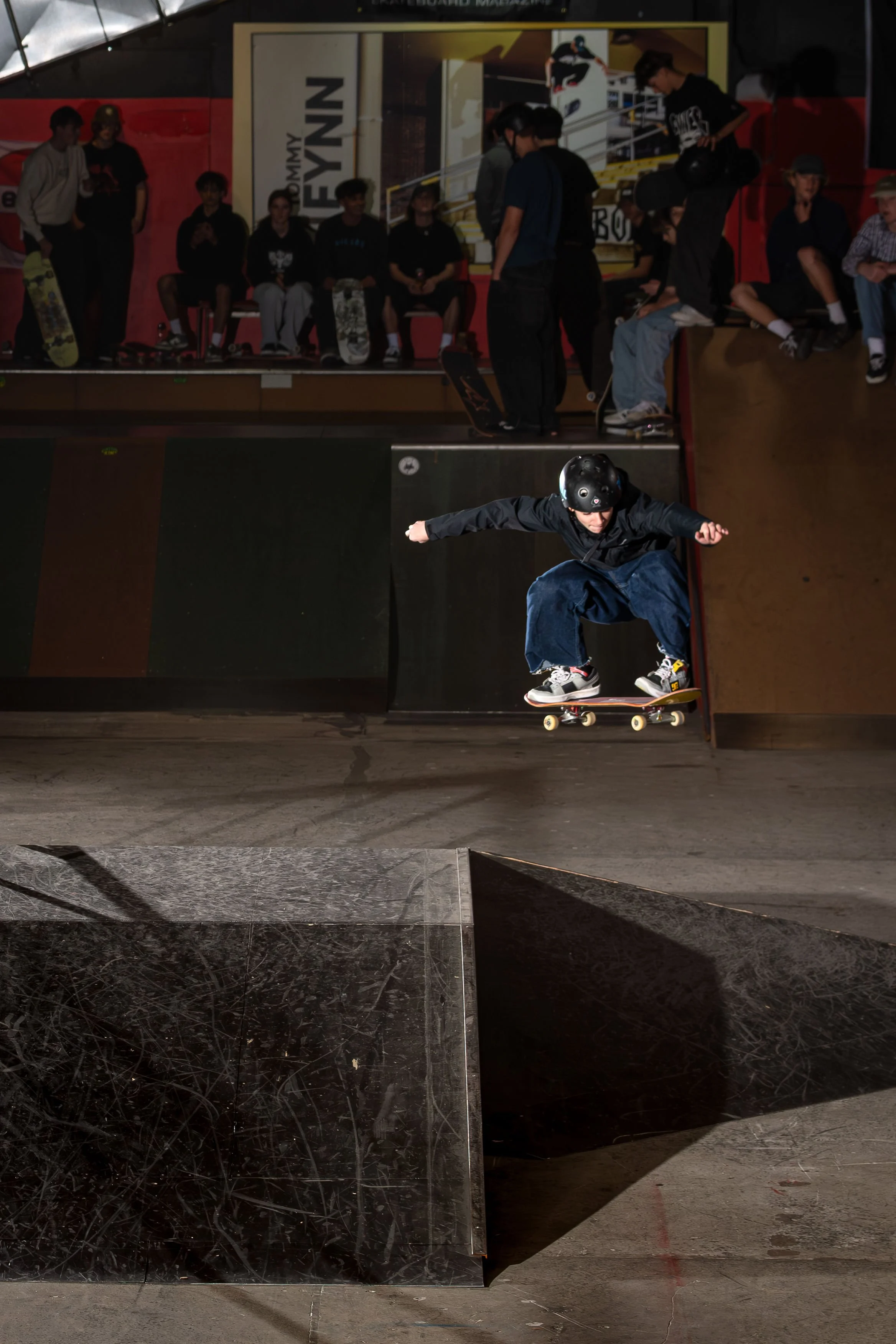 Skateboarder performing a jump in an indoor skate park with an audience watching.