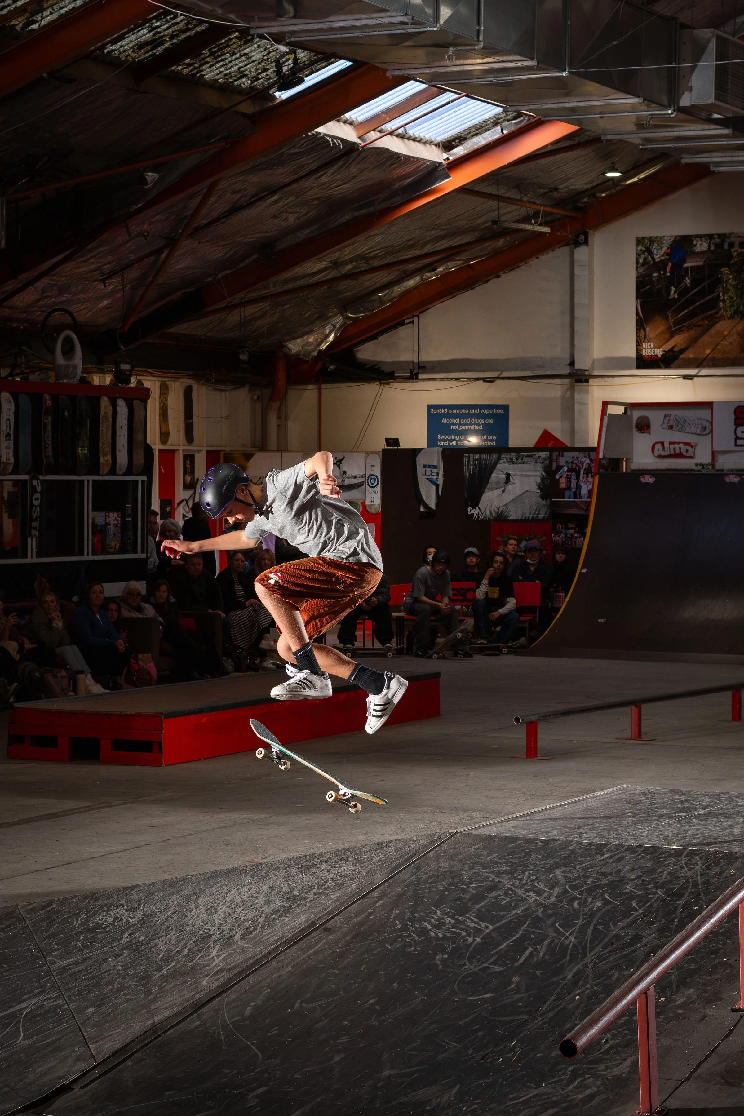 A skateboarder performing a trick indoors at a skate park with ramps and spectators. The individual is in mid-air wearing a helmet, a gray shirt, and shorts.