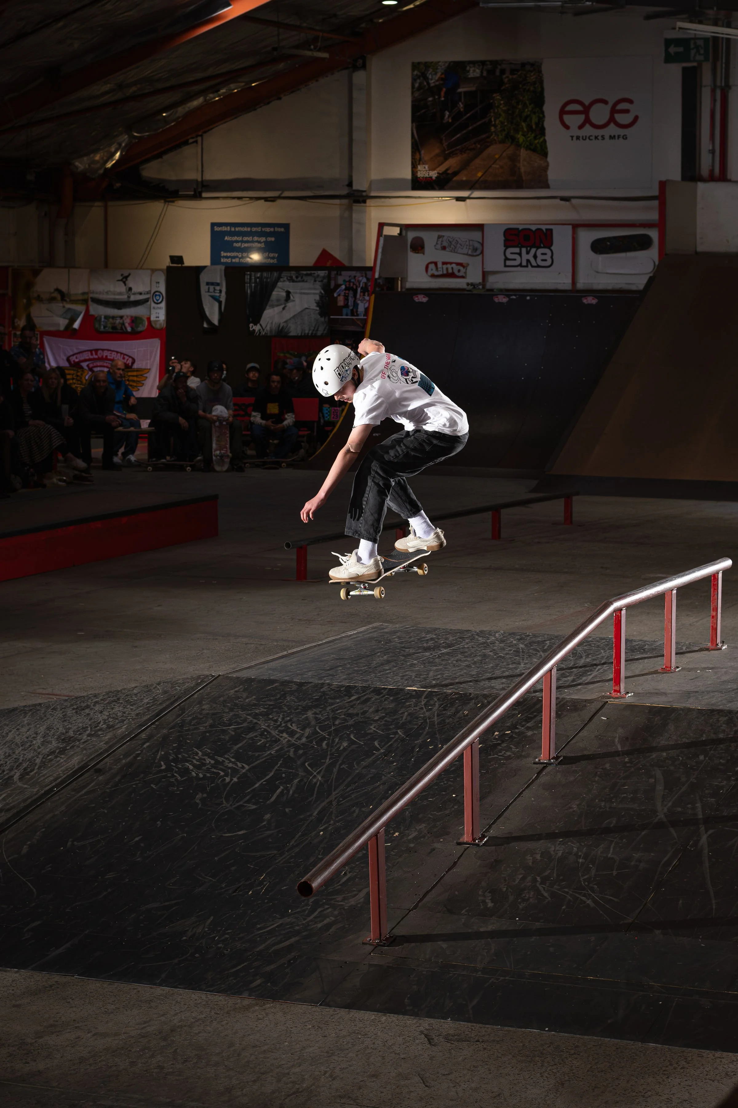 Skateboarder performing a trick in an indoor skate park, wearing a helmet and casual clothing, jumping over a rail. In the background, spectators and skateboarding logos are visible.